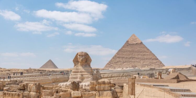 Esfinge de Gizé e pirâmides ao fundo sob céu azul com nuvens esparsas.