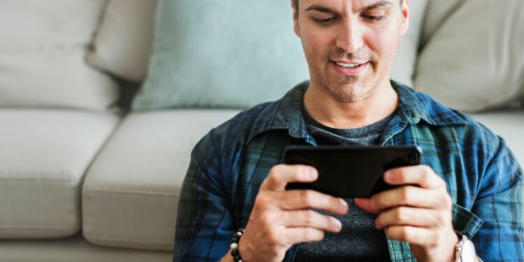 Homem jogando no melhor celular para jogar sentado no sofá segurando smartphone com as duas mãos
