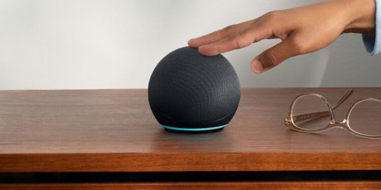 Um Amazon Echo Dot preto esférico com uma faixa de luz azul na base, sobre uma mesa de madeira. Uma mão se aproxima do topo do dispositivo e óculos de leitura estão ao lado.