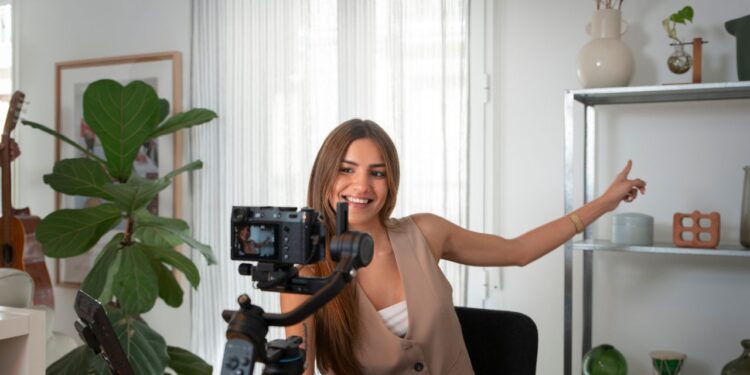 Uma mulher sorridente com longos cabelos castanhos grava um vídeo com uma câmera DSLR montada em um gimbal. Ela veste um colete marrom claro e está sentada em uma mesa de madeira em um quarto bem iluminado com plantas e prateleiras ao fundo, demonstrando como ganhar dinheiro no YouTube pode ser acessível.